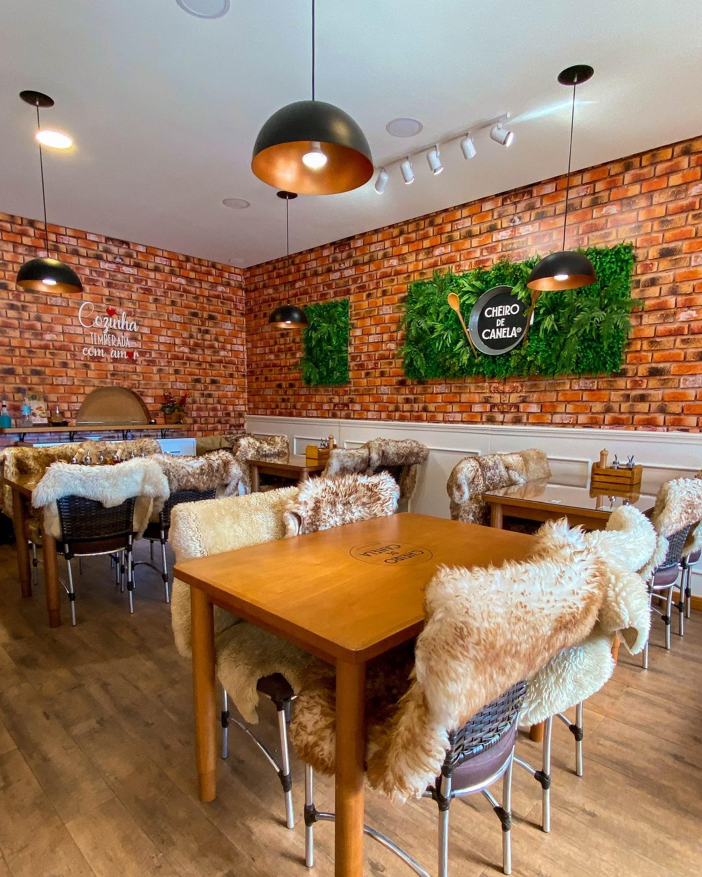 Ambiente interno do Bistrot Cheiro de Canela, com parede de tijolinho e o mural 'Cozinha Temperada com Amor'